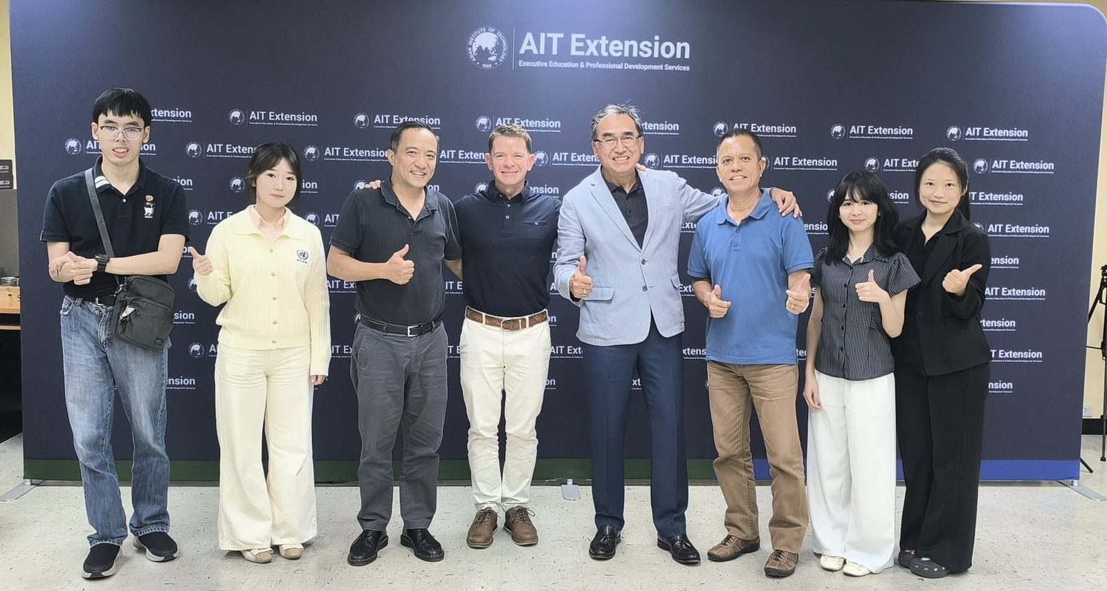 Group photo from Neville Pollard's visit to AIT Extension, in front of the AIT Extension backdrop